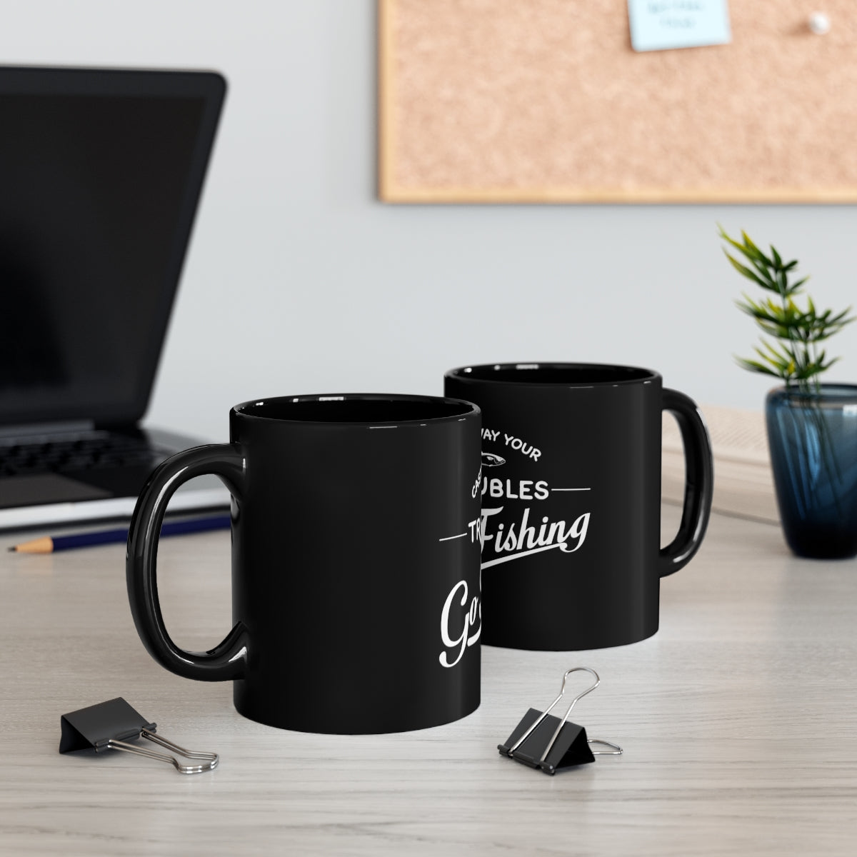 Two black mugs with text on a desk with office supplies and a plant.