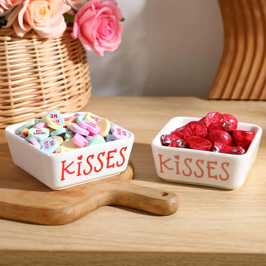 Two ceramic 'Kisses' candy dishes on a wooden surface with flowers in the background.